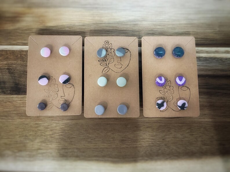 K&ouml;nnte beinhalten: Drei Sets von Ohrsteckern auf braunen Karten. Jede Karte zeigt eine Strichzeichnung eines Gesichts und florale Akzente. Die Ohrringe sind in verschiedenen Farben erh&auml;ltlich, darunter Rosa, Schwarz, Grau, Blau und Lila. Die Ohrringe sind rund.