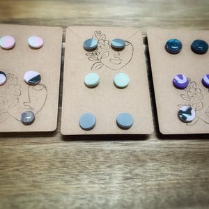 K&ouml;nnte beinhalten: Drei Sets Ohrstecker auf braunen Karten. Jede Karte zeigt ein minimalistisches Gesichtsdesign. Die Ohrringe sind rund und in verschiedenen Farben erh&auml;ltlich, darunter Rosa, Grau, Blau und Lila, einige mit Marmormustern.