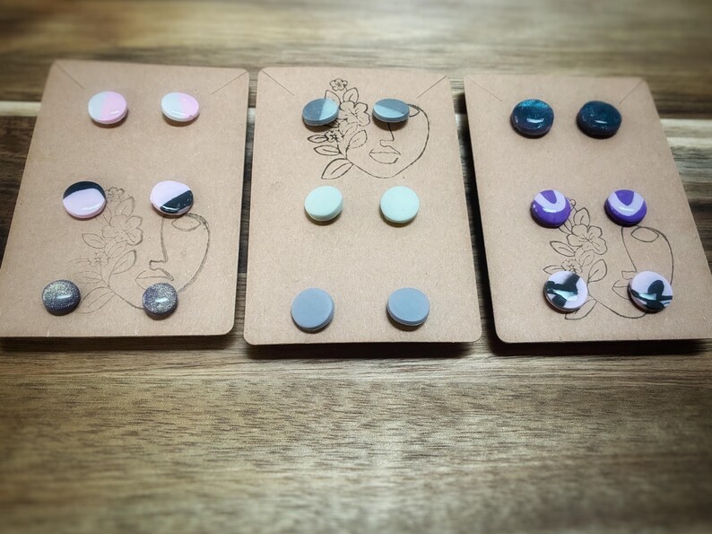 K&ouml;nnte beinhalten: Drei Sets Ohrstecker auf braunen Karten. Jede Karte zeigt ein minimalistisches Gesichtsdesign. Die Ohrringe sind rund und in verschiedenen Farben erh&auml;ltlich, darunter Rosa, Grau, Blau und Lila, einige mit Marmormustern.