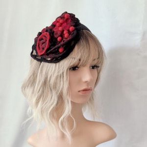 Könnte beinhalten: Ein schwarzes Stirnband mit roten Filzkugeln und einer roten Filzrose. Der Kopfschmuck wird auf einem Schaufensterpuppenkopf mit blonden Haaren präsentiert.