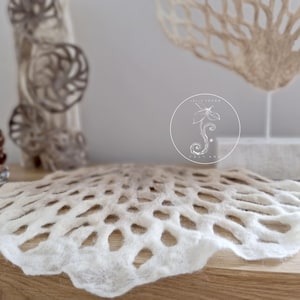 Może przedstawiać: Biało-beżowa wisząca ozdoba ścienna z filcu o delikatnym, koronkowo-podobnym wzorze. Ozdoba ma kształt wachlarza i przechodzi od bieli do beżu.