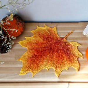 Könnte beinhalten: Ein gefilzter Platzdeckchen in Form eines Ahornblatts mit einem Farbverlauf in warmen Herbsttönen, der von einem tiefroten Zentrum zu leuchtendem Orange und hellgelben Rändern übergeht, mit sichtbaren Faserstrukturen und gezackten Blatträndern.