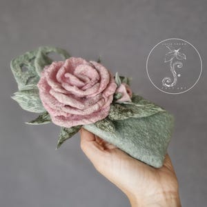 Puede incluir: Tocado floral de fieltro hecho a mano con una gran rosa rosa suave y hojas verdes. El tocado tiene una base verde claro y está acentuado con un capullo rosa más pequeño. El diseño incluye el texto "FELT ART" en un logotipo circular.