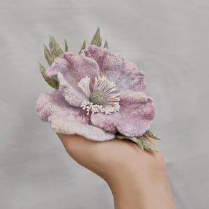 May include: A large, handmade felt flower in shades of lavender and pink, with a detailed white center and green leaves. The flower is held against a light gray background, showcasing its intricate design.