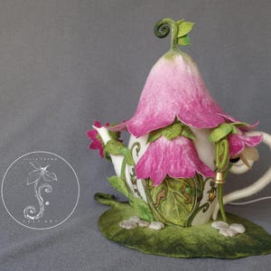Pode incluir: Um bule branco com um design de flores rosa e um padrão de folhas verdes. O bule tem a forma de uma casa com um telhado pontudo e um pequeno caule encaracolado no topo. O bule está sentado em uma base verde em forma de folha.