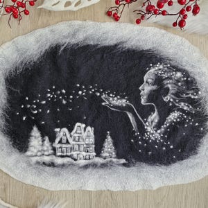 Peut inclure: Une œuvre d'art en laine feutrée représentant une femme soufflant des lumières scintillantes vers un village enneigé. L'œuvre est principalement en noir et blanc, de forme ovale texturée. Des baies rouges et des plumes blanches décorent le fond.