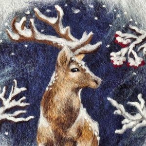 Könnte beinhalten: Ein Filzkunstwerk, das einen Hirsch in einer verschneiten Winterszene darstellt. Der Hirsch hat braunes Fell und ein großes Geweih. Weißer Schnee und Zweige mit roten Beeren umgeben den Hirsch vor einem dunkelblauen Hintergrund.