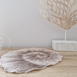 Op de afbeelding: Een beige viltcirkel met een donkerbruin centrum, liggend op een houten oppervlak. Op de achtergrond staat een wit beeld van een koraaltak.