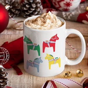 May include: White ceramic mug with a festive design of four colorful Dala horses. The mug is filled with a creamy beverage topped with whipped cream.