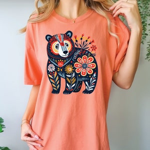 K&ouml;nnte beinhalten: Ein korallenfarbenes T-Shirt mit einem schwarzen B&auml;renmotiv. Der B&auml;r ist von bunten Blumen und Bl&auml;ttern umgeben.