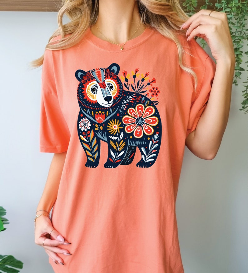 K&ouml;nnte beinhalten: Ein korallenfarbenes T-Shirt mit einem schwarzen B&auml;renmotiv. Der B&auml;r ist von bunten Blumen und Bl&auml;ttern umgeben.