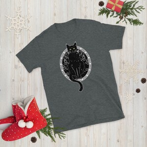 May include: A dark heather grey t-shirt with a black cat sitting inside a circle of stars. The cat has green eyes and the text around the circle reads "The stars are my witness, the moon is my guide, the night is my home".
