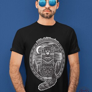 May include: Black t-shirt with a white graphic of a black cat sitting in a forest with a crescent moon and stars. The graphic is surrounded by a circle of text in a stylized font that reads "Julakvittim."