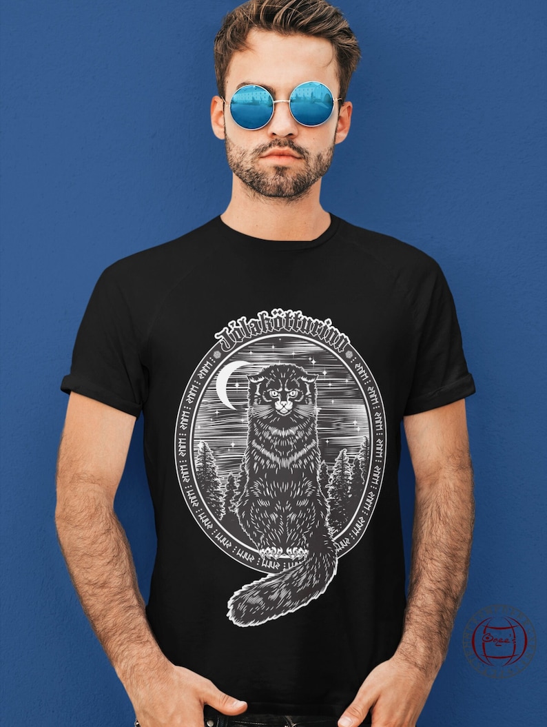 May include: Black t-shirt with a white graphic of a black cat sitting in a forest with a crescent moon and stars. The graphic is surrounded by a circle of text in a stylized font that reads "Julakvittim."