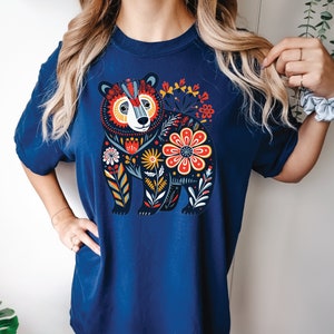 K&ouml;nnte beinhalten: Ein marineblaues T-Shirt mit einem farbenfrohen Blumendesign, das einen B&auml;ren zeigt. Der B&auml;r ist schwarz umrandet und mit verschiedenen Farben gef&uuml;llt, darunter Rot, Gelb, Blau und Gr&uuml;n. Die Blumen sind ebenfalls bunt und haben verschiedene Formen und Gr&ouml;&szlig;en.