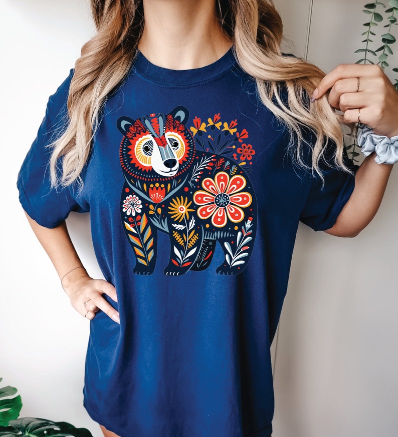 K&ouml;nnte beinhalten: Ein marineblaues T-Shirt mit einem farbenfrohen Blumendesign, das einen B&auml;ren zeigt. Der B&auml;r ist schwarz umrandet und mit verschiedenen Farben gef&uuml;llt, darunter Rot, Gelb, Blau und Gr&uuml;n. Die Blumen sind ebenfalls bunt und haben verschiedene Formen und Gr&ouml;&szlig;en.