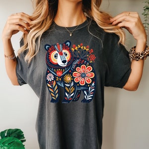 K&ouml;nnte beinhalten: Ein dunkelgraues T-Shirt mit einem farbenfrohen Blumendesign, das einen B&auml;ren zeigt. Der B&auml;r ist von Blumen in Rot-, Gelb-, Blau- und Gr&uuml;nt&ouml;nen umgeben.