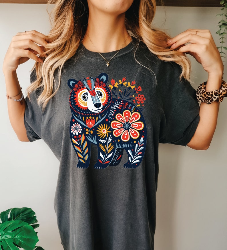 K&ouml;nnte beinhalten: Ein dunkelgraues T-Shirt mit einem farbenfrohen Blumendesign, das einen B&auml;ren zeigt. Der B&auml;r ist von Blumen in Rot-, Gelb-, Blau- und Gr&uuml;nt&ouml;nen umgeben.