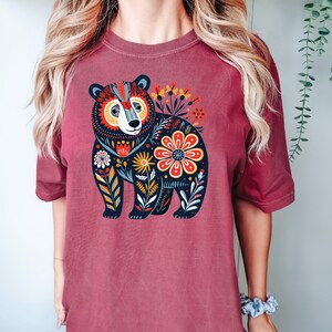 K&ouml;nnte beinhalten: Ein maroonfarbenes T-Shirt mit einer Illustration eines schwarzen B&auml;ren. Der B&auml;r ist von bunten Blumen und Bl&auml;ttern umgeben. Das Design ist im Stil der Volkskunst.