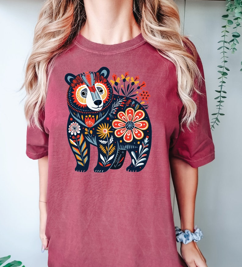 K&ouml;nnte beinhalten: Ein maroonfarbenes T-Shirt mit einer Illustration eines schwarzen B&auml;ren. Der B&auml;r ist von bunten Blumen und Bl&auml;ttern umgeben. Das Design ist im Stil der Volkskunst.