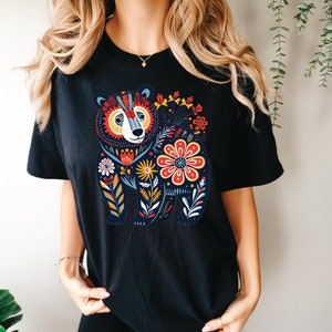 K&ouml;nnte beinhalten: Schwarzes T-Shirt mit einem farbenfrohen Blumendesign, das einen B&auml;ren zeigt. Der B&auml;r ist von Blumen in Rot-, Gelb-, Blau- und Gr&uuml;nt&ouml;nen umgeben.