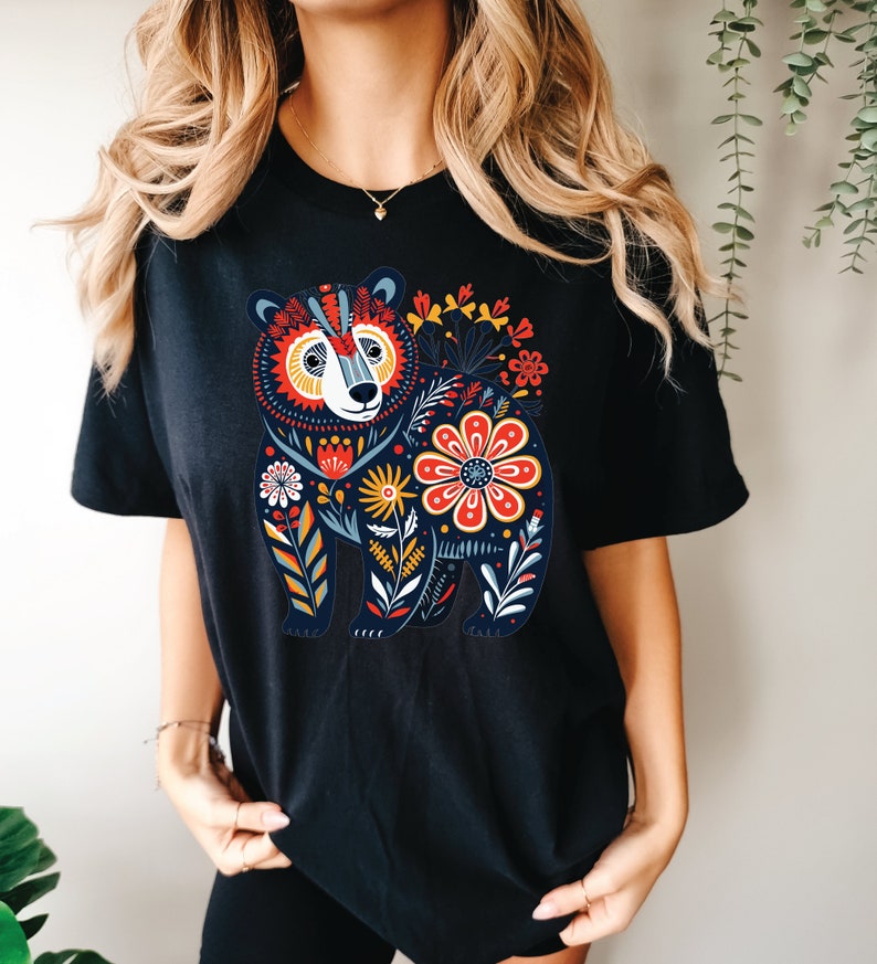 K&ouml;nnte beinhalten: Schwarzes T-Shirt mit einem farbenfrohen Blumendesign, das einen B&auml;ren zeigt. Der B&auml;r ist von Blumen in Rot-, Gelb-, Blau- und Gr&uuml;nt&ouml;nen umgeben.