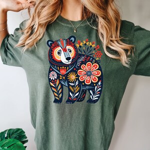 K&ouml;nnte beinhalten: Ein gr&uuml;nes T-Shirt mit einer bunten Illustration eines B&auml;ren, umgeben von Blumen. Der B&auml;r ist schwarz mit wei&szlig;en Akzenten und die Blumen sind rot, gelb, orange und blau.