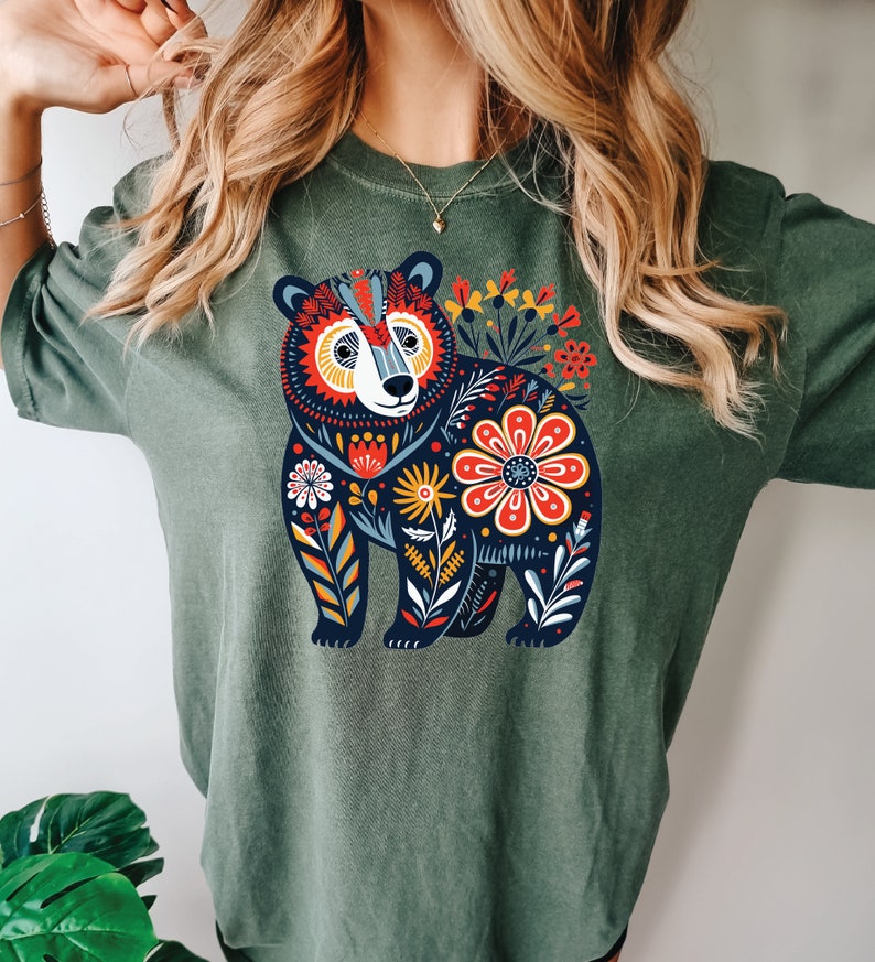 K&ouml;nnte beinhalten: Ein gr&uuml;nes T-Shirt mit einer bunten Illustration eines B&auml;ren, umgeben von Blumen. Der B&auml;r ist schwarz mit wei&szlig;en Akzenten und die Blumen sind rot, gelb, orange und blau.