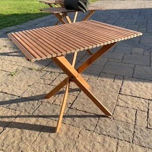 Può includere: Un tavolo pieghevole in legno con un piano a doghe e una base a croce. Il tavolo è realizzato in legno naturale e ha un aspetto rustico.