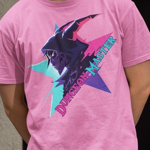 Puede incluir: Una camiseta rosa con un gráfico de una figura encapuchada con cuernos y el texto "Dungeon Master" en una fuente colorida y estilizada.