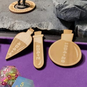 May include: A tabletop role-playing game setup with miniature figures, dice, and wooden potion bottle tokens. The scene includes a purple tray, a rocky terrain, and various dice.