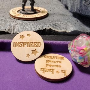 May include: A tabletop gaming scene featuring dice and wooden tokens. One token reads "INSPIRED" with star cutouts. Another reads "GREATER HEALTH POTION". A miniature figure stands on a wooden base. Dice are translucent with colorful inclusions.