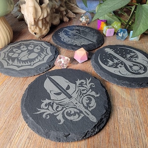 May include: Four black slate coasters with different designs. The designs include a paw print, a sword, a lute, and a floral pattern. The coasters are on a wooden surface with dice and other decorative items.