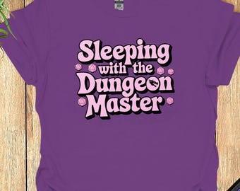 Może przedstawiać: Fioletowy T-shirt z napisem "Sleeping with the Dungeon Master" w różowych literach z różowymi kostkami do gry wokół tekstu.