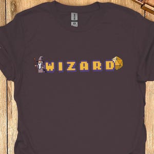 May include: A dark purple t-shirt with a pixelated design of a wizard holding a staff and a yellow and orange dice. The text "WIZARD" is in yellow with purple outlines.
