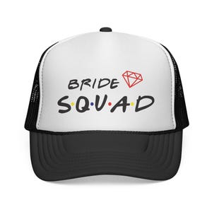 May include: A black and white trucker hat featuring a white front panel with the black text BRIDE SQUAD. The letters are separated by small colored dots, and a red diamond outline is positioned to the right of the word BRIDE.