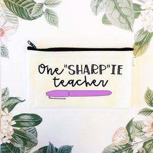 Puede incluir: Estuche de lápices color crema con cremallera negra y el texto "One "SHARP"IE teacher" en escritura negra. Un gráfico de marcador Sharpie morado está debajo del texto. El estuche es rectangular y se encuentra sobre una superficie blanca con detalles florales.