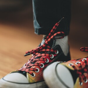 Pu&ograve; includere: Primo piano di un paio di scarpe da ginnastica nere e rosse a scacchi con suole bianche. Le scarpe hanno un design a fiamma sul lato e sono legate con lacci rossi e neri.