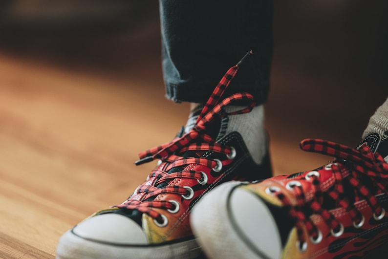 Pu&ograve; includere: Primo piano di un paio di scarpe da ginnastica nere e rosse a scacchi con suole bianche. Le scarpe hanno un design a fiamma sul lato e sono legate con lacci rossi e neri.
