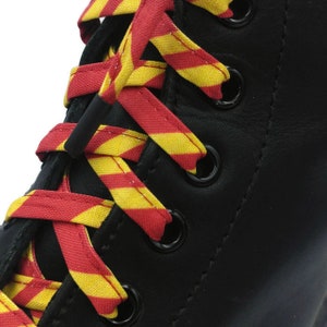 Puede incluir: Botas de cuero negro con cordones rayados rojos y amarillos. Los cordones están atados en forma de cruz.