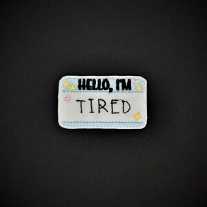 Peut inclure: Étiquette de nom en feutre blanc avec une bordure bleue et des étoiles et des cœurs brodés colorés. L'étiquette indique "Hello, I'm Tired".