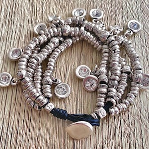 May include: Silver bracelet with multiple strands of small, round beads. The bracelet has dangling, circular charms, each set with a clear, sparkling stone. The bracelet is fastened with a black cord and a silver clasp. A stylish piece of jewellery.