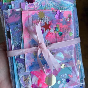 May include: A stack of rectangular paper cards featuring abstract pink, blue, and purple paint strokes, tied with a light pink ribbon. The cards include printed text about zodiac signs and gemstones, decorated with small metallic star and heart sequins.