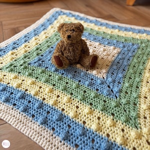 May include: A small brown teddy bear sits on a handmade, square crochet blanket. The blanket features concentric stripes of cream, light blue, yellow, and green, with a textured, bobble stitch pattern. The bear has brown fur and dark brown paws.