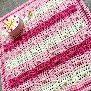 May include: A pink and white striped crochet baby blanket with a textured stitch pattern. The blanket has a white border and is made with soft yarn.