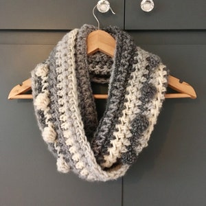 May include: A grey and white crocheted infinity scarf with a textured bobble stitch pattern. The scarf is hanging on a wooden hanger.