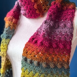 May include: A colorful crocheted scarf with a gradient of yellow, green, brown, orange, pink, and red. The scarf is made with a textured stitch that creates a bumpy surface.