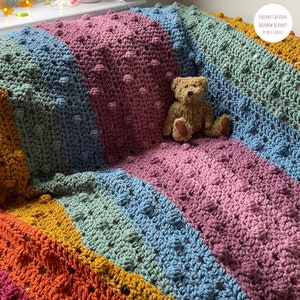 May include: A colorful crocheted blanket with a chevron pattern and textured bobbles. The blanket is draped over a couch with a teddy bear sitting on top. The blanket is made in a rainbow of colors, including red, orange, yellow, green, blue, and purple.