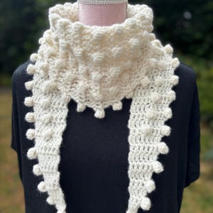 May include: A cream-coloured crochet scarf with a textured bobble stitch design. The scarf, displayed on a mannequin, has a wide collar and long, fringed ends. This handmade scarf is a cosy accessory.