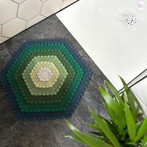 hexagon shaped bath mat made with crocheted cotton yarn using bobble stitch and tr crochet in tones of green. shown in place in a shower room with a plant